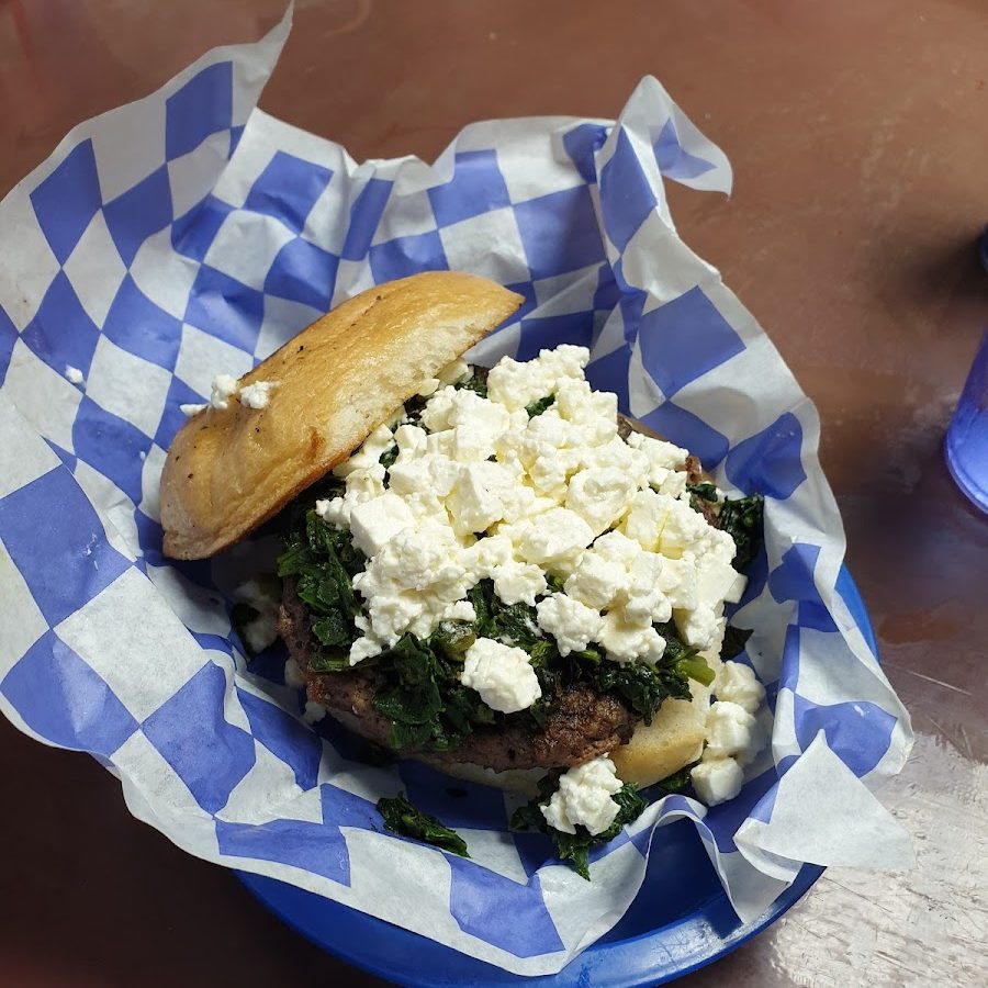 An 8oz Greek burger topped with spinach and crumbled feta cheese in a blue checkered basket